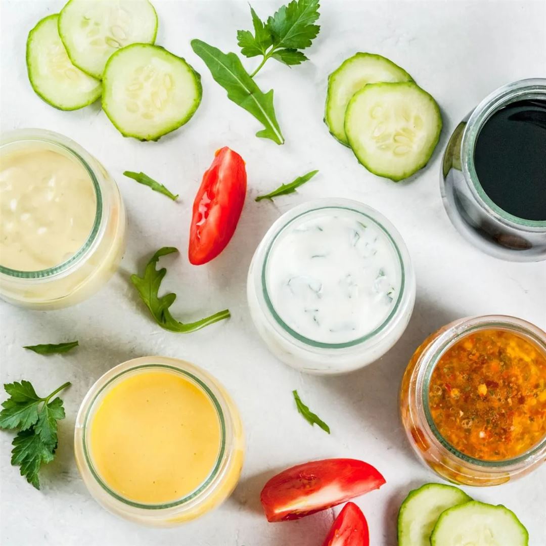 Five jars of different salad dressings are arranged on a white surface, surrounded by cucumber slices, tomato wedges, arugula, and parsley for a fresh and colorful presentation.